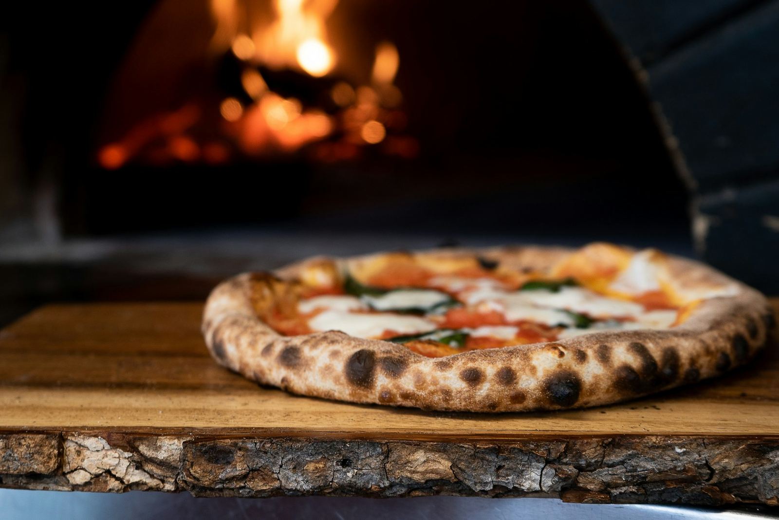 Pizza margherita appena sfornata davanti alla bocca di un forno a legna acceso